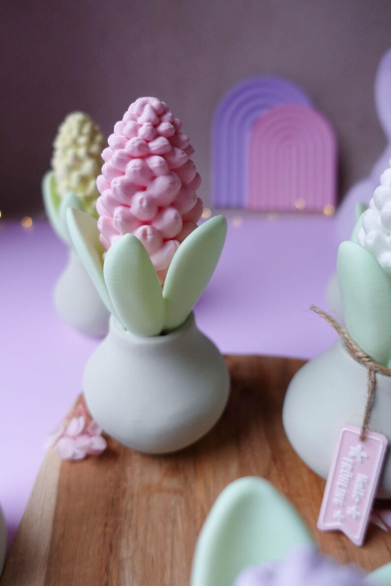May include: A collection of decorative floral arrangements. Each arrangement features a pastel-colored flower, with a pink, yellow, and white flower, set in a white vase with green leaves. A wooden board and a purple background complete the scene.