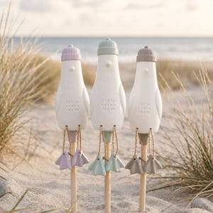 Könnte beinhalten: Drei weiße Keramikvogel-Figuren mit glatten Körpern und texturierten Brustdetails. Jeder Vogel trägt eine einfarbige Mütze in Lavendel, Salbeigrün oder Taupe. Sie haben baumelnde Keramik-Schwimmfüße, die mit Schnur an Holzstäben befestigt sind.