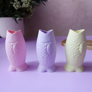 Puede incluir: Tres jarrones de cerámica con forma de pez en colores pastel: rosa, lavanda y amarillo pálido. Cada jarrón tiene un patrón de escamas texturizadas y una base en forma de aleta. Los jarrones se exhiben sobre una superficie púrpura.