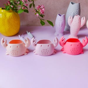 Könnte beinhalten: Drei krabbenförmige Tee-Eier in Rosatönen mit orangefarbenen Innenräumen und kleinen Löchern. Im Hintergrund eine gelbe Vase mit Grün und rosa Blüten, sowie fisch- und hummerförmige Vasen. Die Artikel befinden sich auf einer lila Oberfläche.