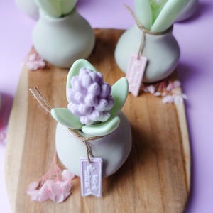 May include: A wooden serving board displays several small, decorative vases. Each vase holds a sculpted flower, some in pink, white, and lavender. The vases are light green, and each has a small tag tied with twine. The background is a soft purple.