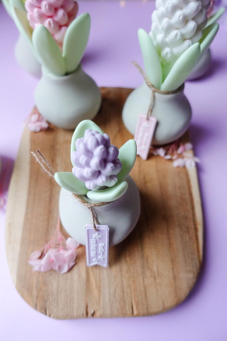 May include: A wooden serving board displays several small, decorative vases. Each vase holds a sculpted flower, some in pink, white, and lavender. The vases are light green, and each has a small tag tied with twine. The background is a soft purple.