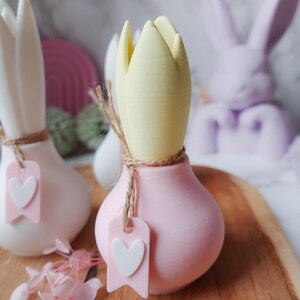 K&ouml;nnte beinhalten: Nahaufnahme von dekorativen tulpenf&ouml;rmigen Keramikvasen in Wei&szlig; und Rosa, mit einer gelben Tulpenknospe. Jede Vase hat eine Schnur und ein rosa Etikett mit einem wei&szlig;en Herzen. Im Hintergrund eine Lavendel-Hasenfigur.