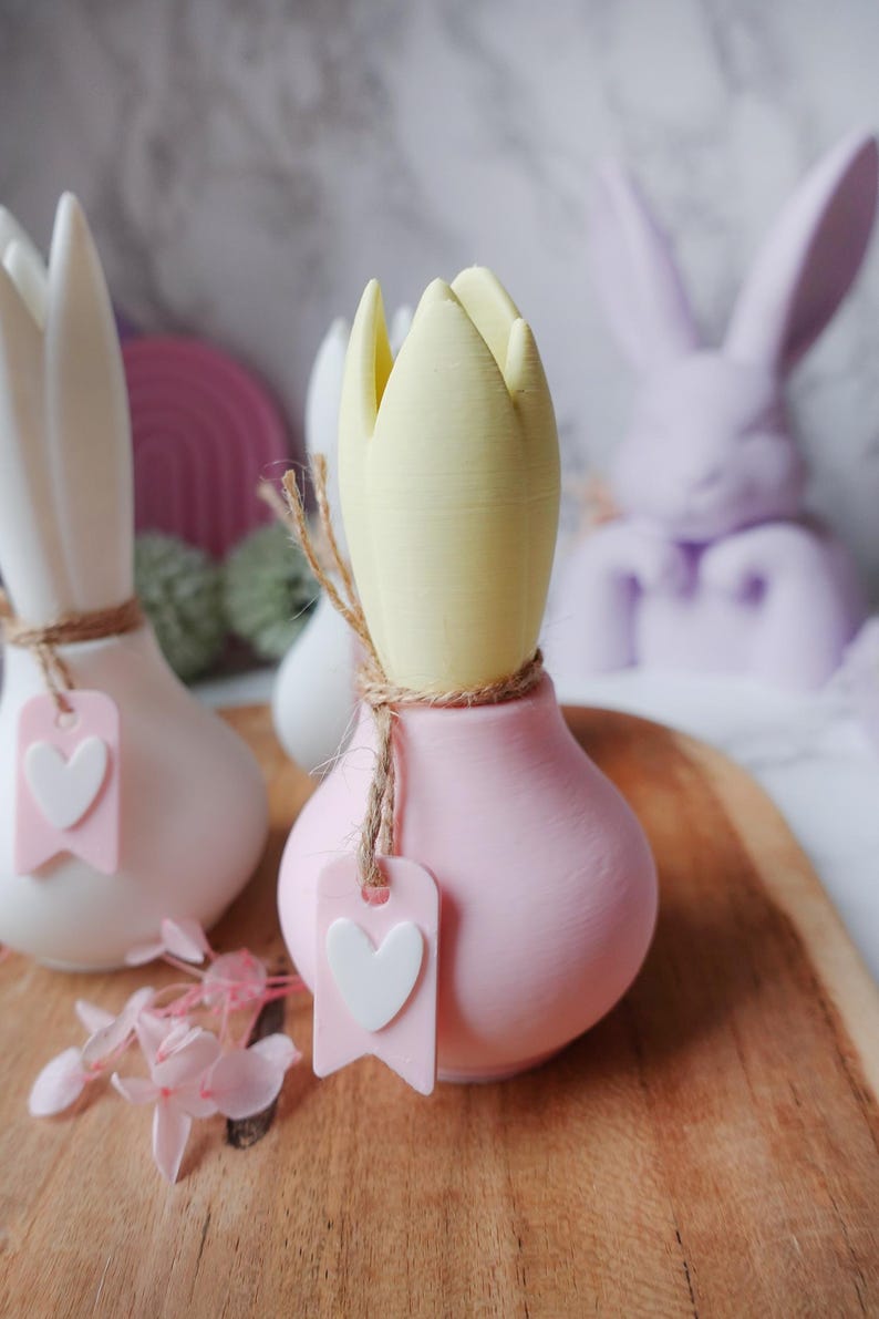 K&ouml;nnte beinhalten: Nahaufnahme von dekorativen tulpenf&ouml;rmigen Keramikvasen in Wei&szlig; und Rosa, mit einer gelben Tulpenknospe. Jede Vase hat eine Schnur und ein rosa Etikett mit einem wei&szlig;en Herzen. Im Hintergrund eine Lavendel-Hasenfigur.
