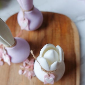 K&ouml;nnte beinhalten: Nahaufnahme von drei dekorativen tulpenf&ouml;rmigen Objekten. Eines ist wei&szlig;, zwei sind lavendelfarben. Jedes hat eine Schnur und ein kleines Etikett mit einem Herzen. Die Objekte befinden sich auf einer Holzoberfl&auml;che mit verstreuten rosa floralen Akzenten.