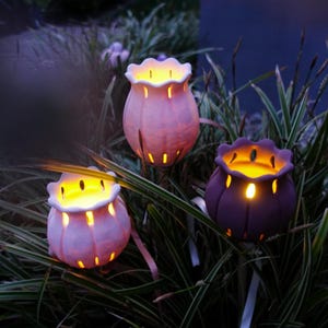 May include: Three illuminated, tulip-shaped garden lights in varying shades of purple and pink. The lights are set among green foliage, with a soft glow emanating from within. The lights are on white stakes.