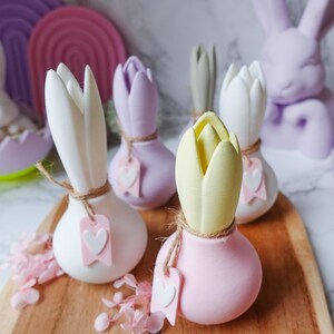 K&ouml;nnte beinhalten: Eine Sammlung pastellfarbener tulpenf&ouml;rmiger Dekorationen auf einem Holzbrett. Die Tulpen sind wei&szlig;, lavendelfarben, gelb und rosa, jede mit einer Schnur und einem herzf&ouml;rmigen Anh&auml;nger. Im Hintergrund befindet sich eine Lavendel-Hasenstatue.