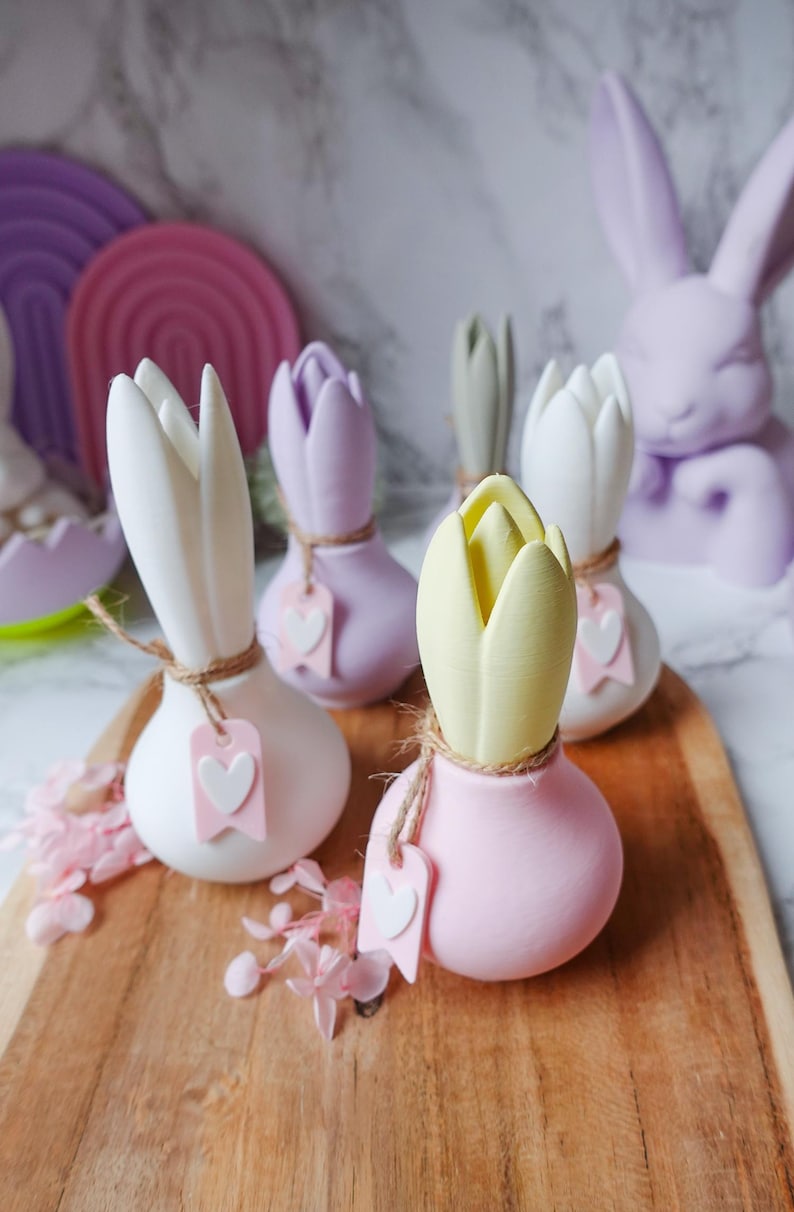 K&ouml;nnte beinhalten: Eine Sammlung pastellfarbener tulpenf&ouml;rmiger Dekorationen auf einem Holzbrett. Die Tulpen sind wei&szlig;, lavendelfarben, gelb und rosa, jede mit einer Schnur und einem herzf&ouml;rmigen Anh&auml;nger. Im Hintergrund befindet sich eine Lavendel-Hasenstatue.