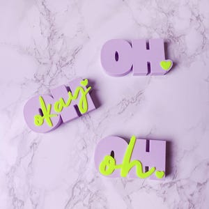 Può includere: Tre elementi di word art 3D color lavanda con le parole "OH" e "okay" in un font giocoso. La parola "okay" è in verde neon, e la parola "OH" ha un cuore di accento verde neon. I pezzi sono su una superficie di marmo.