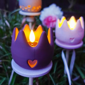 Op de afbeelding: Drie kroonvormige kaarsenhouders op stokjes, één donkerpaars en twee lichtroze, elk voorzien van een hartvormige uitsparing en een gloeiende led-vlam. De houders zijn gemonteerd op ronde plateaus bovenop dunne stelen en stralen warm licht uit.