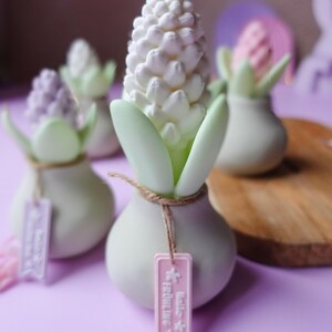 May include: A collection of decorative hyacinth-shaped objects. Each piece features a white, textured flower head, green leaves, and a small tag. The objects are set on a purple surface, with a wooden board in the background.