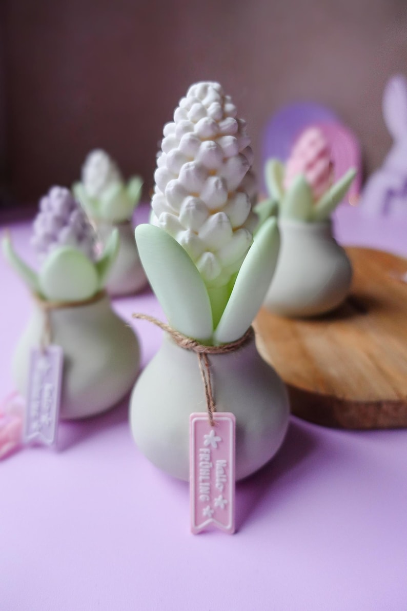 May include: A collection of decorative hyacinth-shaped objects. Each piece features a white, textured flower head, green leaves, and a small tag. The objects are set on a purple surface, with a wooden board in the background.