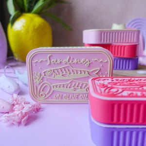 Puede incluir: Contenedores rosas en forma de sardinas con el texto "Sardines in Olive Oil". Los contenedores están apilados en varios colores, incluyendo rosa, rojo y morado. Un jarrón amarillo con flores está en el fondo.