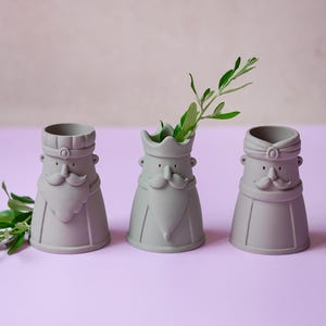 以下が含まれることがあります： 口ひげとあごひげを生やした様式化された人物の形をした、マットグレーの陶器製容器3点。それぞれの容器には王冠やターバンなど、異なる頭飾りが施されている。1つの容器には小さな緑の葉の小枝が入っている。
