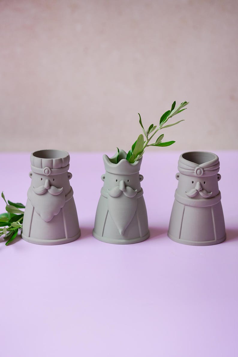 Peut inclure: Trois r&eacute;cipients en c&eacute;ramique gris mat en forme de personnages stylis&eacute;s avec moustaches et barbes. Chaque r&eacute;cipient pr&eacute;sente un couvre-chef distinct, incluant une couronne et des turbans. Un r&eacute;cipient contient un petit brin de feuilles vertes.