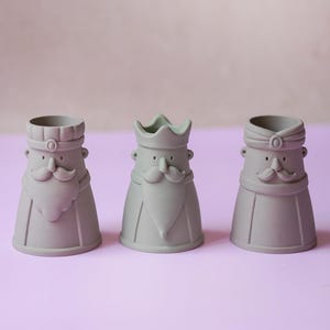 Peut inclure: Trois tasses en c&eacute;ramique gris mat en forme de personnages stylis&eacute;s avec moustaches et barbes. Chaque tasse pr&eacute;sente un couvre-chef distinct : une casquette &agrave; sommet plat, une couronne &agrave; trois pointes et un turban. Visages simplifi&eacute;s.