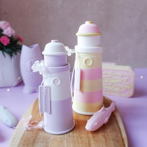 Könnte beinhalten: Zwei dekorative Leuchtturm-förmige Behälter in Pastellfarben, einer lavendelfarben und weiß, der andere gelb und rosa gestreift. Eine kleine rosa Fischfigur und ein Holztablett sind ebenfalls vorhanden. Die Leuchttürme haben ein Etikett mit "Kinderliebe".
