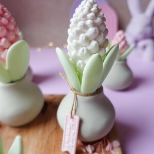 May include: A white hyacinth flower sculpture with green leaves in a small, round, light green vase. A pink tag hangs from the vase. Other similar sculptures are in the background, on a wooden surface, with a purple background.