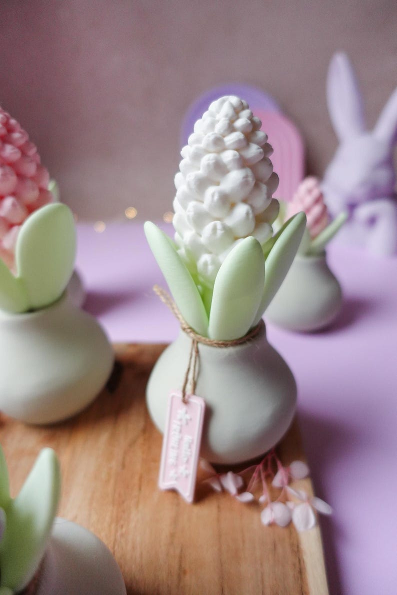May include: A white hyacinth flower sculpture with green leaves in a small, round, light green vase. A pink tag hangs from the vase. Other similar sculptures are in the background, on a wooden surface, with a purple background.