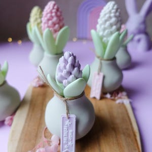 May include: A collection of decorative hyacinth flower sculptures in pastel colors, including pink, white, and purple, set in small, light gray vases with green leaves. Each vase has a tag that says "Hello Fr&uuml;hling". The arrangement is on a wooden board.