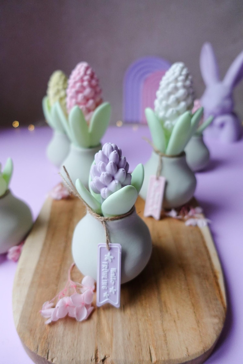 May include: A collection of decorative hyacinth flower sculptures in pastel colors, including pink, white, and purple, set in small, light gray vases with green leaves. Each vase has a tag that says "Hello Fr&uuml;hling". The arrangement is on a wooden board.
