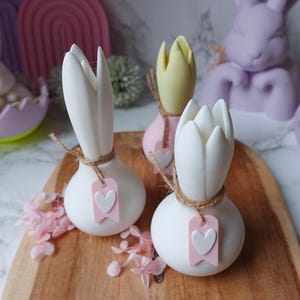 K&ouml;nnte beinhalten: Wei&szlig;e, tulpenf&ouml;rmige Dekorationsartikel mit Juteband und einem rosa Herzanh&auml;nger. Auf einem Holzbrett, mit einem hellrosa und gelben tulpenf&ouml;rmigen Artikel im Hintergrund. Osterdekoration.