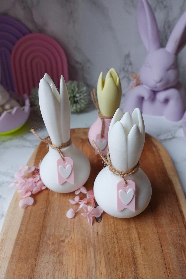 K&ouml;nnte beinhalten: Wei&szlig;e, tulpenf&ouml;rmige Dekorationsartikel mit Juteband und einem rosa Herzanh&auml;nger. Auf einem Holzbrett, mit einem hellrosa und gelben tulpenf&ouml;rmigen Artikel im Hintergrund. Osterdekoration.