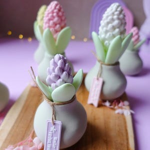 May include: A collection of decorative flower-shaped objects in pastel colors. Each piece features a bulbous flower head in pink, white, or purple, set in a small, light green vase with matching leaves. Some have tags that read "Hello Fr&uuml;hlings".