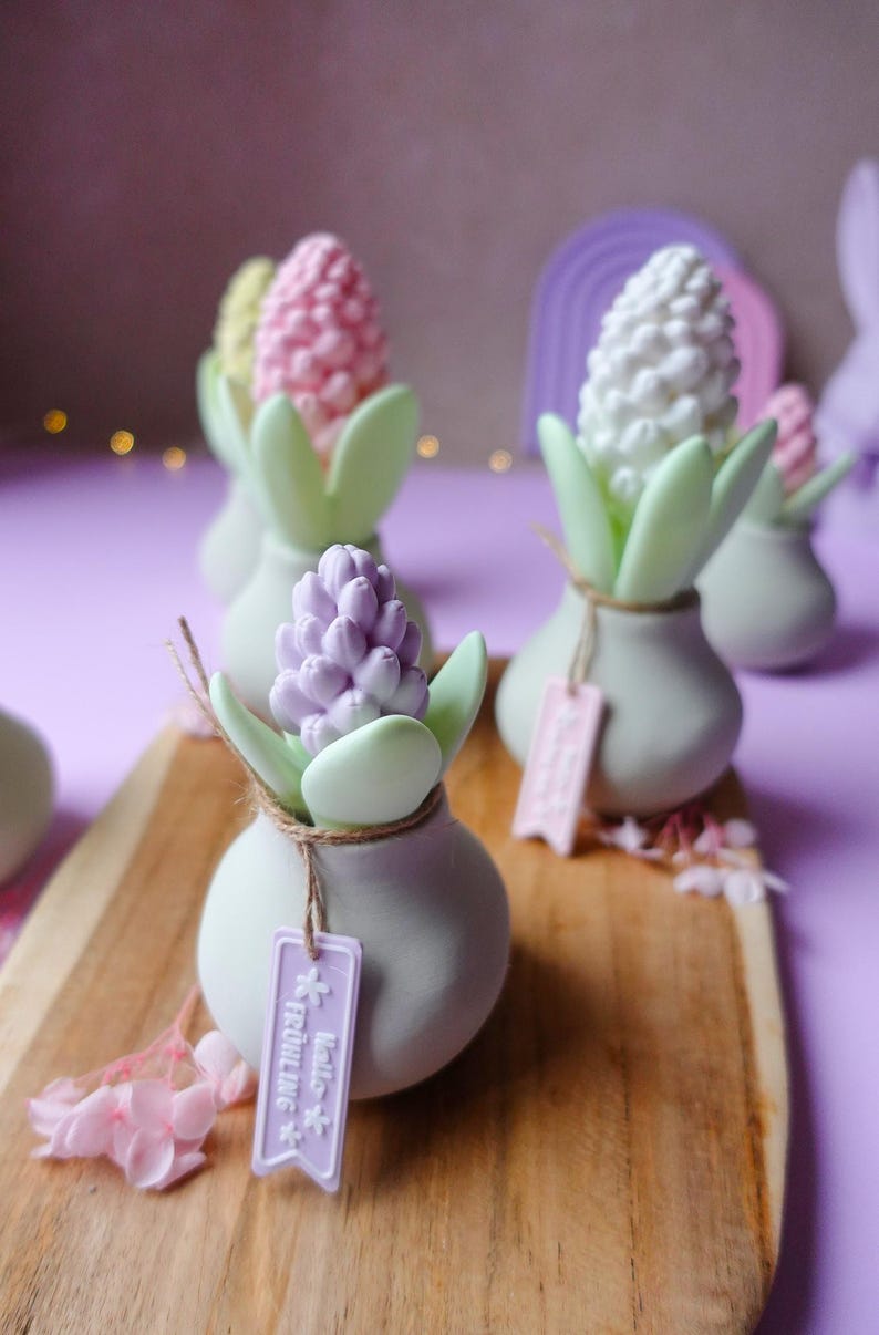 May include: A collection of decorative flower-shaped objects in pastel colors. Each piece features a bulbous flower head in pink, white, or purple, set in a small, light green vase with matching leaves. Some have tags that read "Hello Fr&uuml;hlings".