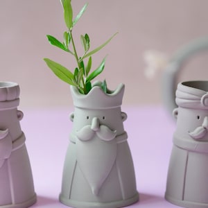 Peut inclure: Trois vases en c&eacute;ramique gris clair en forme de personnages avec couronnes, moustaches et barbes. Le vase central contient une petite branche verte feuillue. Chaque r&eacute;cipient pr&eacute;sente une finition mate lisse et une base large et stable.