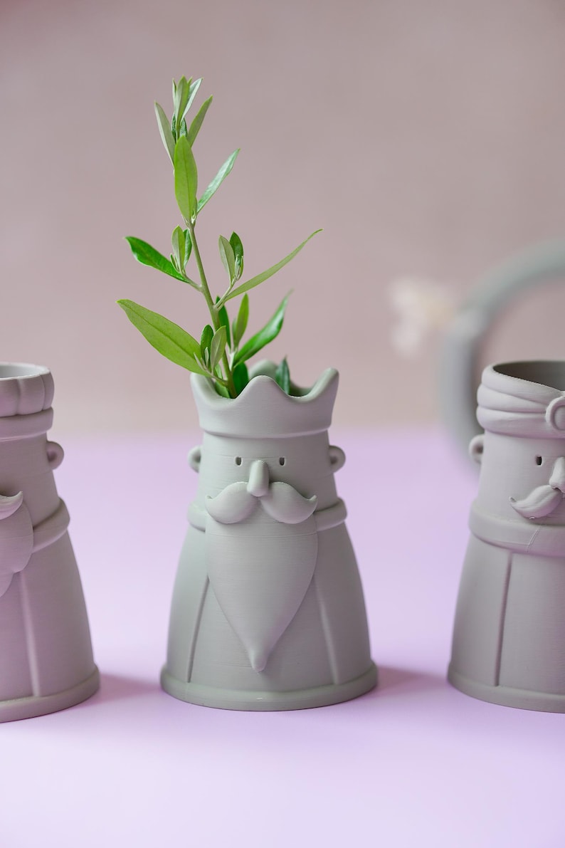 Peut inclure: Trois vases en c&eacute;ramique gris clair en forme de personnages avec couronnes, moustaches et barbes. Le vase central contient une petite branche verte feuillue. Chaque r&eacute;cipient pr&eacute;sente une finition mate lisse et une base large et stable.