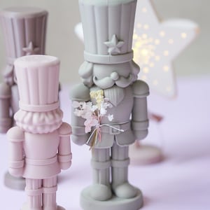 May include: Three nutcracker figurines in matte shades of gray, dusty pink, and mauve. The central gray nutcracker features a tall hat with a star emblem, a mustache, and a small bouquet of dried white and pink flowers tied to its chest.
