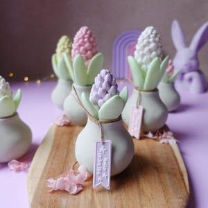Op de afbeelding: Een verzameling decoratieve hyacintbloemsculpturen in pastelkleuren, gerangschikt op een houten plank. Elke sculptuur staat in een kleine, gebroken witte vaas met groene bladeren en een label met "Hello Frühling". Paarse achtergrond.
