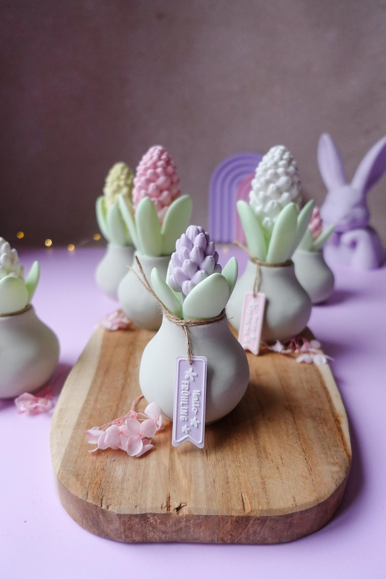 May include: A collection of decorative hyacinth flower sculptures in pastel colors, arranged on a wooden board. Each sculpture is in a small, off-white vase with green leaves and a tag that says "Hello Fr&uuml;hling". A purple background complements the display.