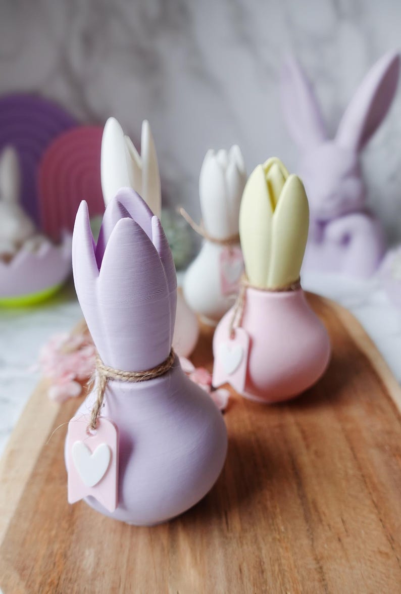 K&ouml;nnte beinhalten: Eine Sammlung von tulpenf&ouml;rmigen Dekorationen in Pastellfarben. Die Tulpen sind in den Farbt&ouml;nen Lavendel, Wei&szlig; und Gelb gehalten, mit einer Schnurakzentuierung und einem herzf&ouml;rmigen Anh&auml;nger. Ein Holztablett und Osterhasen-Dekorationen befinden sich im Hintergrund.
