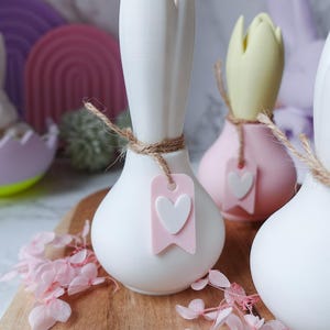 K&ouml;nnte beinhalten: Wei&szlig;e, rosa und gelbe dekorative Vasen in Zwiebelform mit einem herzf&ouml;rmigen Anh&auml;nger, der mit Bindfaden befestigt ist. Die Vasen stehen auf einem Holzbrett mit rosa Bl&uuml;ten. Der Hintergrund umfasst pastellfarbene dekorative Formen.