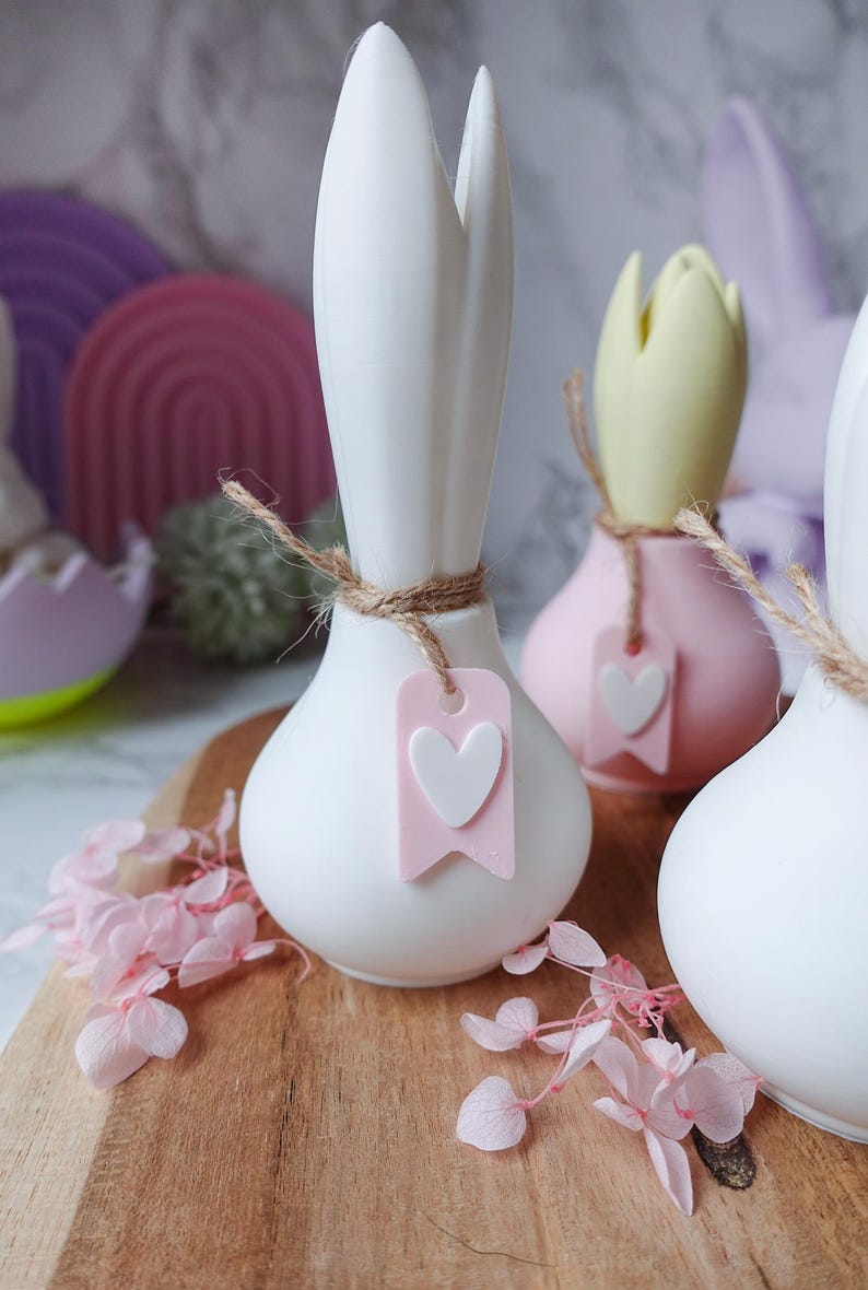 K&ouml;nnte beinhalten: Wei&szlig;e, rosa und gelbe dekorative Vasen in Zwiebelform mit einem herzf&ouml;rmigen Anh&auml;nger, der mit Bindfaden befestigt ist. Die Vasen stehen auf einem Holzbrett mit rosa Bl&uuml;ten. Der Hintergrund umfasst pastellfarbene dekorative Formen.