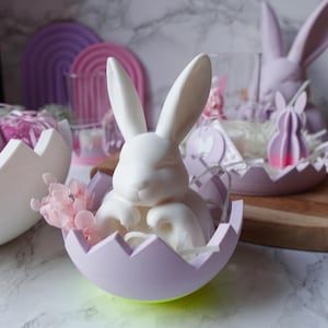 May include: A white bunny figurine nestled in a lavender eggshell, accented with pink flowers. Other Easter-themed decorations include a pink bunny, candles, and rainbow-shaped decor. The items are arranged on a marble surface.