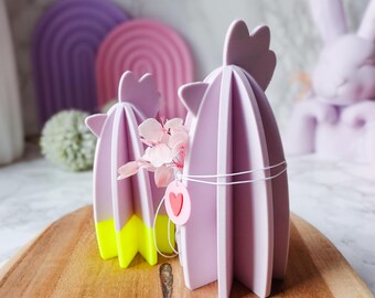 May include: Two lavender cactus-shaped decorations with yellow accents and a pink heart tag, displayed on a wooden tray. The decorations are part of a collection of pastel-colored Easter-themed decor.