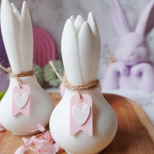 K&ouml;nnte beinhalten: Zwei wei&szlig;e Keramikvasen in Tulpenform mit Juteschnur und einem rosa Anh&auml;nger mit wei&szlig;em Herz. Im Hintergrund eine Lavendel-Hasenfigur. Die Vasen stehen auf einer Holzoberfl&auml;che, mit rosa Blumen und einem rosa Regenbogen im Hintergrund.