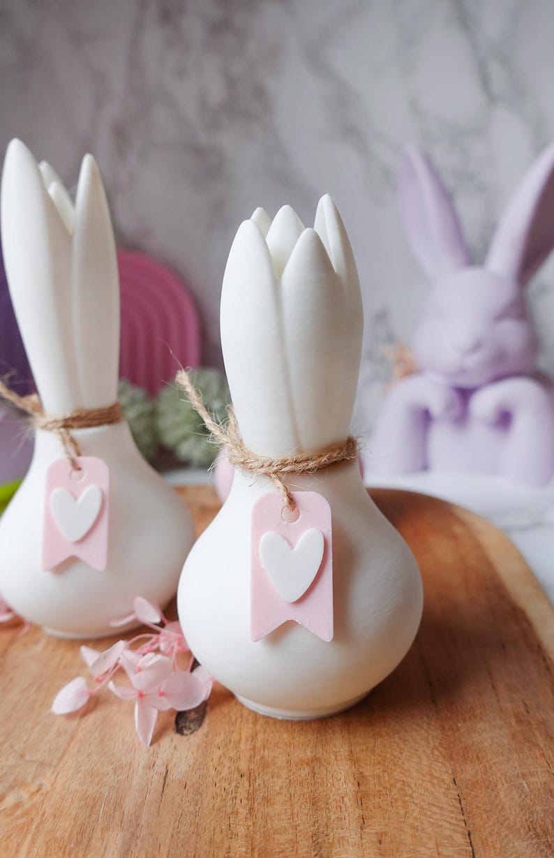 K&ouml;nnte beinhalten: Zwei wei&szlig;e Keramikvasen in Tulpenform mit Juteschnur und einem rosa Anh&auml;nger mit wei&szlig;em Herz. Im Hintergrund eine Lavendel-Hasenfigur. Die Vasen stehen auf einer Holzoberfl&auml;che, mit rosa Blumen und einem rosa Regenbogen im Hintergrund.