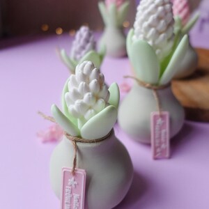 May include: Several small, white ceramic vases with hyacinth-shaped decorations. Each vase has a light green stem and leaves, with a white or pink flower. The vases are tied with twine and have pink tags that read "Hello Fr&uuml;hling."