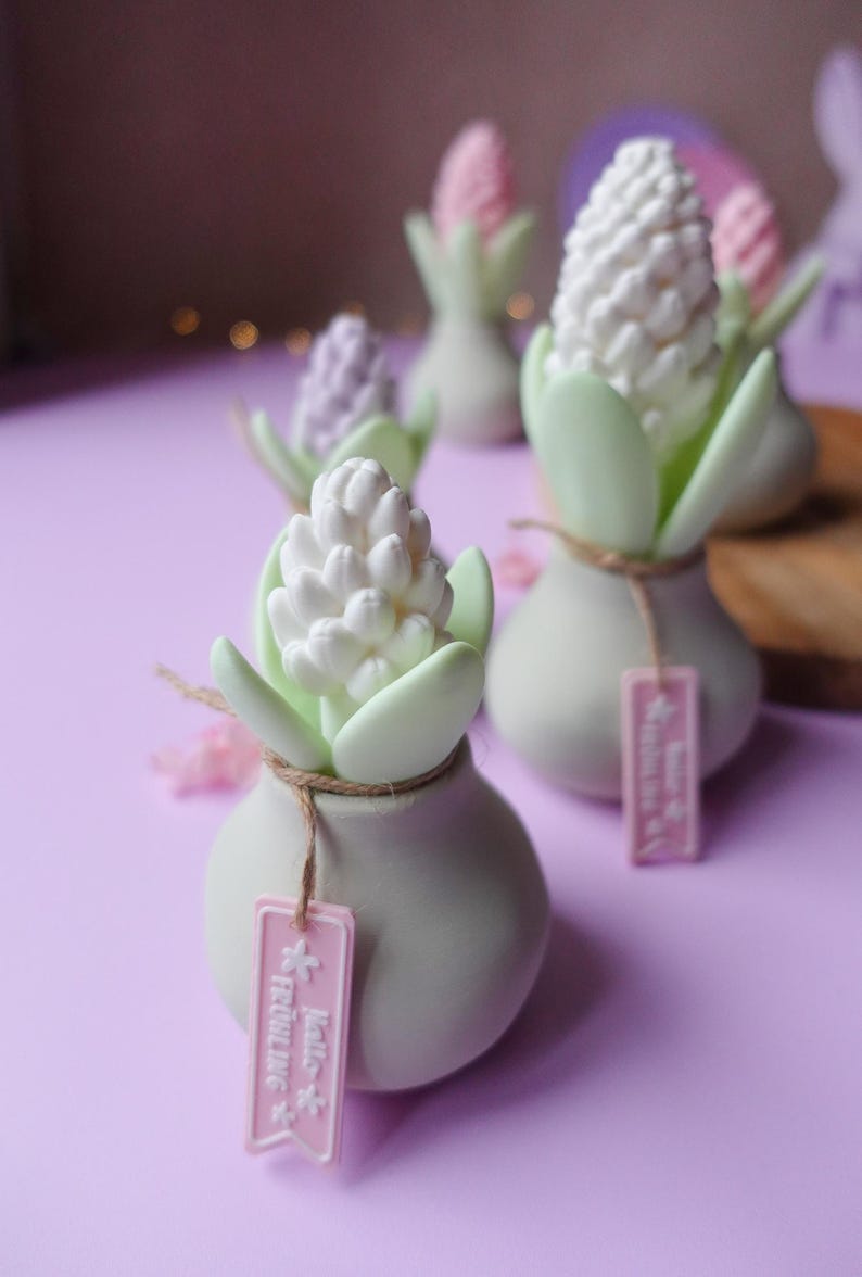 May include: Several small, white ceramic vases with hyacinth-shaped decorations. Each vase has a light green stem and leaves, with a white or pink flower. The vases are tied with twine and have pink tags that read "Hello Fr&uuml;hling."