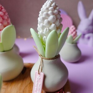 May include: A white hyacinth flower sculpture with green leaves in a small, light gray vase. A pink tag hangs from the vase. Other similar sculptures are in the background, with a wooden surface and a purple background.