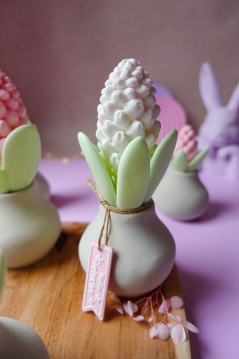 May include: A white hyacinth flower sculpture with green leaves in a small, light gray vase. A pink tag hangs from the vase. Other similar sculptures are in the background, with a wooden surface and a purple background.