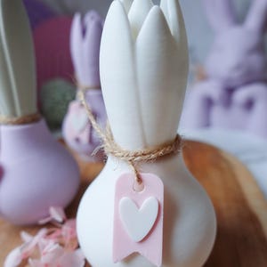 K&ouml;nnte beinhalten: Wei&szlig;e tulpenf&ouml;rmige Keramikvase mit Jute-Akzent und einem rosa Anh&auml;nger mit wei&szlig;em Herz. Weitere pastellfarbene Hasenvasen im Hintergrund. Die Vasen stehen auf einer Holzoberfl&auml;che.