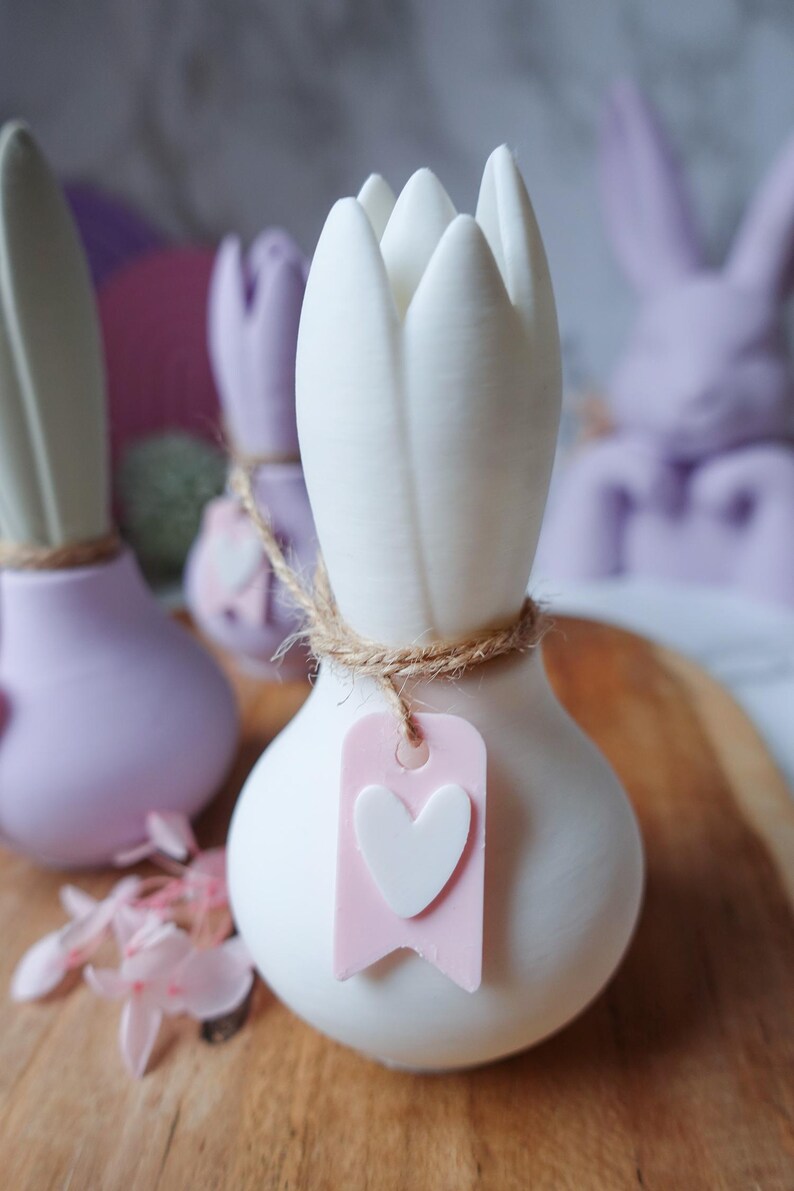 K&ouml;nnte beinhalten: Wei&szlig;e tulpenf&ouml;rmige Keramikvase mit Jute-Akzent und einem rosa Anh&auml;nger mit wei&szlig;em Herz. Weitere pastellfarbene Hasenvasen im Hintergrund. Die Vasen stehen auf einer Holzoberfl&auml;che.