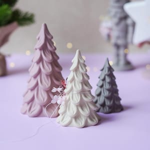 以下が含まれることがあります： サイズの異なる3つの装飾用ツリーフィギュアのセット。一番大きいものはモーブ色、中くらいのものはクリーム色で小さな花のアクセントがあり、一番小さいものはダークグレーです。それぞれのツリーの表面には、花びらが重なったような層状のテクスチャーが施されています。