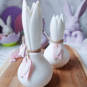 K&ouml;nnte beinhalten: Wei&szlig;e, tulpenf&ouml;rmige Keramikvasen mit Schnurakzenten und rosa Herzanh&auml;ngern auf einem Holzbrett. Die Vasen sind Teil einer Osterdekoration mit pastellfarbenen Hasenfiguren und dekorativen Eiern im Hintergrund. Die Vasen sind ca. 15 cm hoch.
