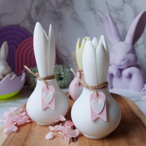 Puede incluir: Jarrones decorativos de cerámica blanca con diseño de tulipán, atados con cuerda y adornados con etiquetas rosas en forma de corazón. Tema de Pascua con decoraciones pastel.